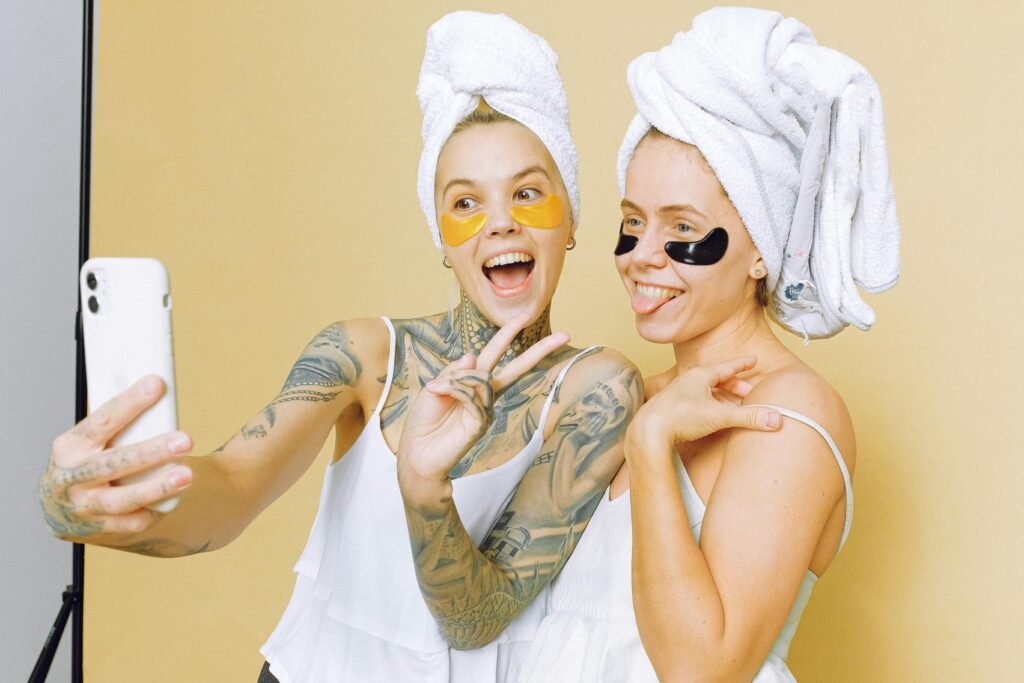 Two women enjoying a skincare routine with facial masks, taking a selfie wrapped in towels against a beige background.