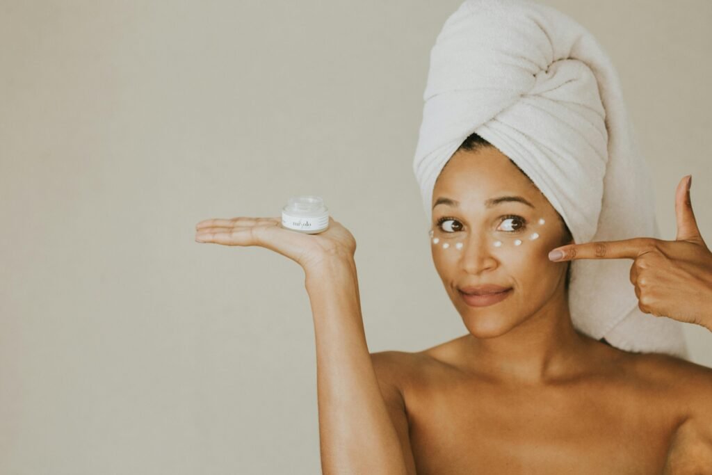 A woman applies under eye cream, holding a jar, with a towel on her head.