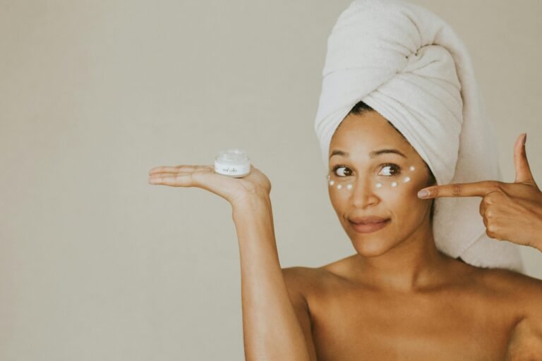 A woman applies under eye cream, holding a jar, with a towel on her head.