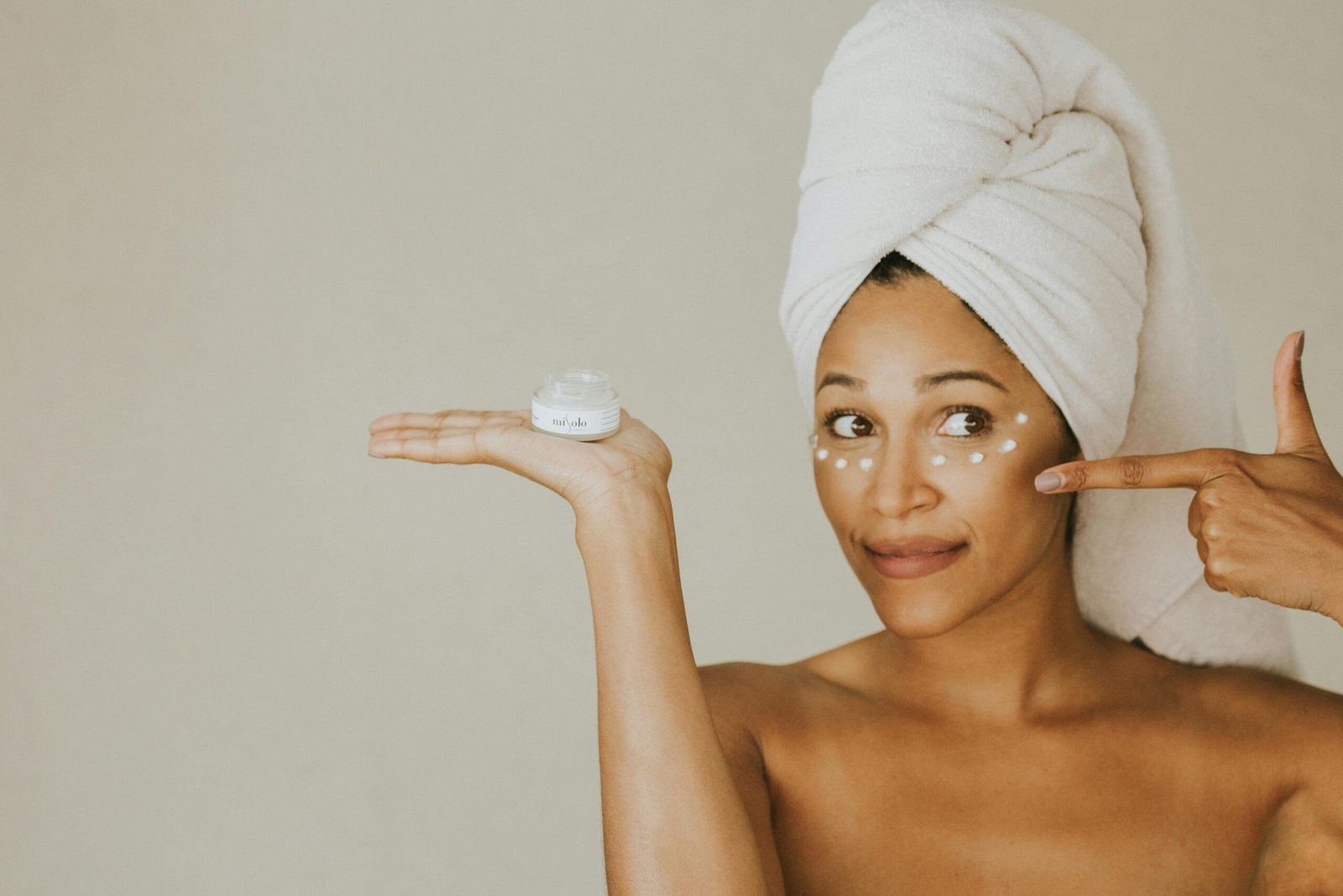 A woman applies under eye cream, holding a jar, with a towel on her head.